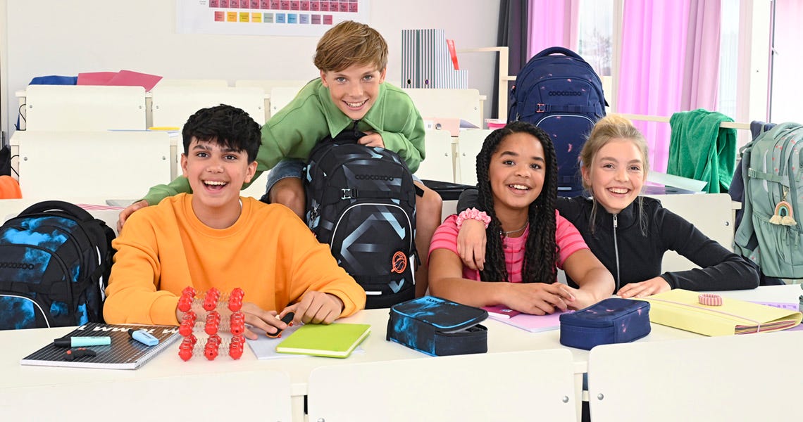 Vier Freunde sitzen lächelnd im Klassenzimmer mit ihren coocazoo Schulrucksäcken