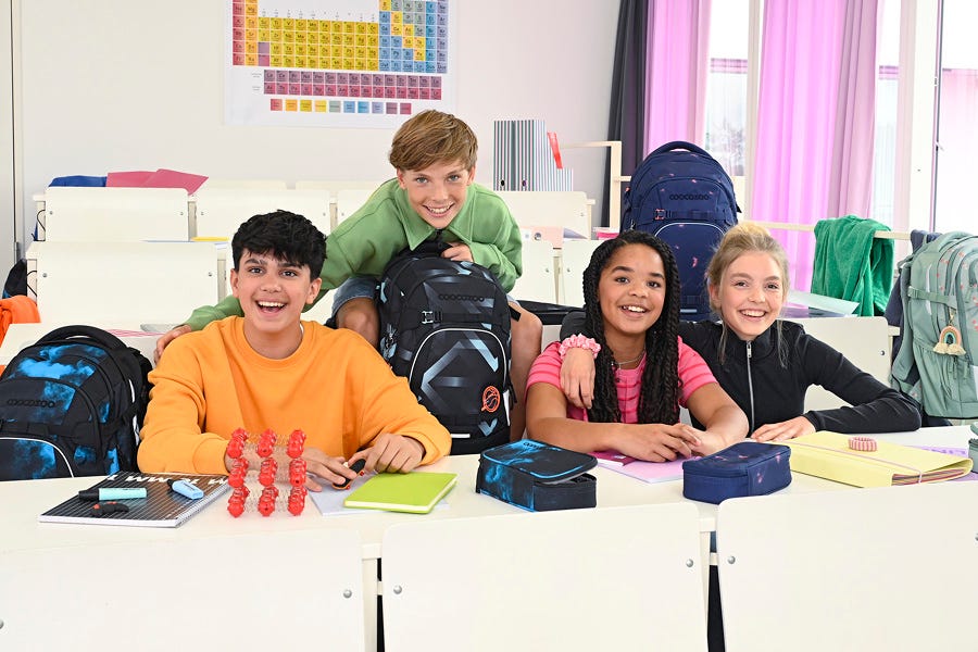 Vier Freunde sitzen lächelnd im Klassenzimmer mit ihren coocazoo Schulrucksäcken