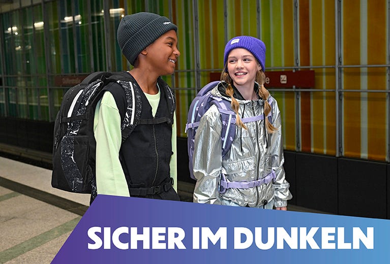 Zwei Kinder mit reflektierenden Elementen am Schulrucksack laufen an einer U-Bahn Station entlang